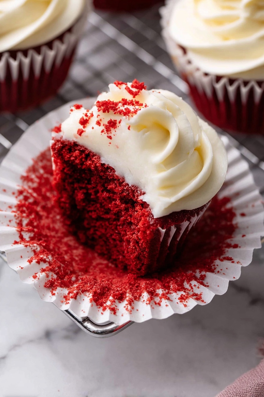 A single red velvet cupcake sits in an opened white paper liner that is stained with bright red crumbs. The cupcake has one layer of deep red, moist, and dense cake topped with a swirl of creamy white frosting that has a smooth, slightly fluffy texture. Some red crumbs are lightly sprinkled on top of the frosting, adding contrast. The cupcake is placed on a white marbled surface with a metal cooling rack partially visible beneath it. In the background, there are parts of other similarly frosted cupcakes. photo taken with an iphone --ar 2:3 --v 7