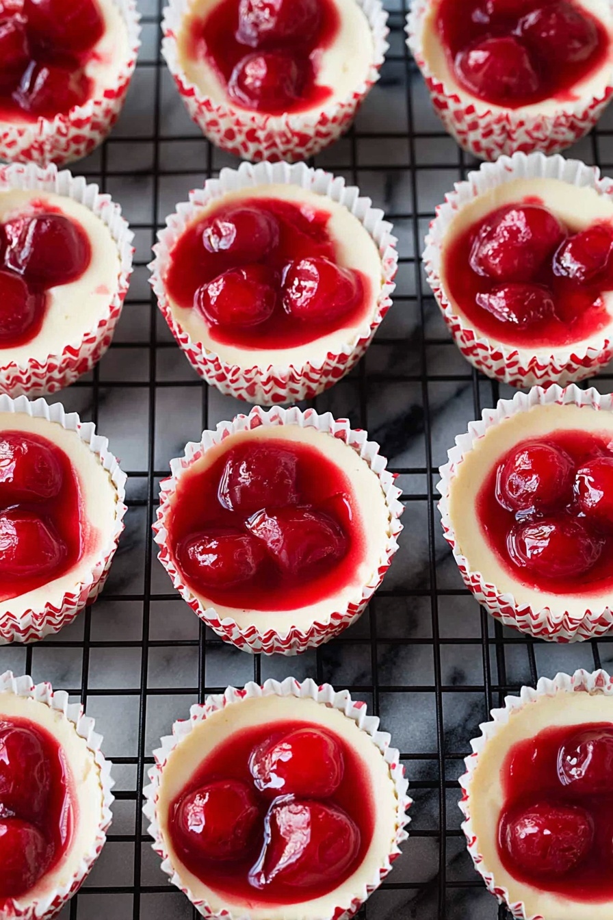 The image shows many small cheesecakes in white paper cups with red patterns, arranged neatly on a black wire cooling rack over a white marbled surface. Each cheesecake has two clear layers: the bottom layer is creamy white and smooth, filling most of the cup, while the top layer is a bright, glossy red cherry topping with whole cherries visible inside. The cherry topping is slightly uneven and shiny, peeking over the edge of the creamy base, and the paper cups create a textured, ruffled border around each cheesecake. photo taken with an iphone --ar 2:3 --v 7