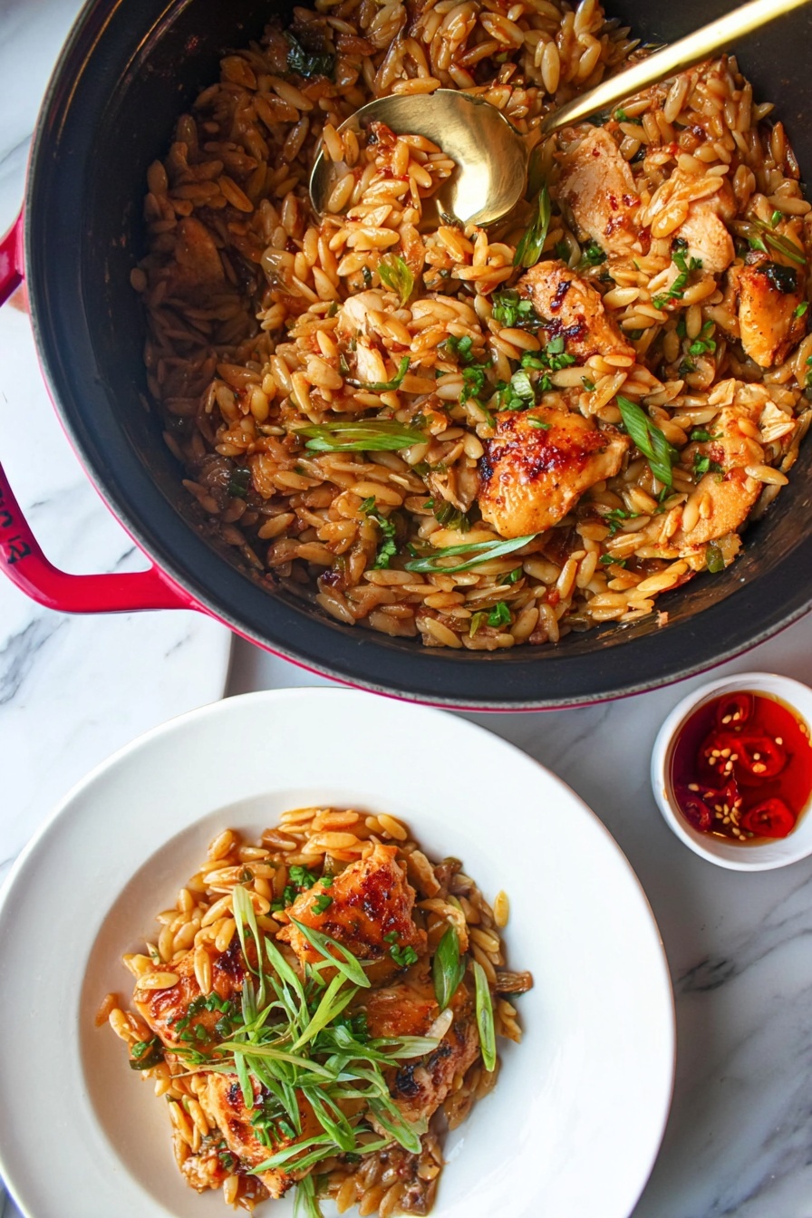 A close-up image shows a black pot with red handles filled with a dish made of small oval-shaped toasted rice pasta mixed with several pieces of cooked golden-brown chicken, garnished with green herbs and thin slices of spring onions. A gold spoon is in the pot lifting some of the food. Below the pot, a white plate holds a serving of the same dish with a few green herbs on top. To the right of the plate, there is a small white bowl with a red-colored sauce that has a visible chili pepper inside. The whole scene is set on a white marbled surface. photo taken with an iphone --ar 2:3 --v 7