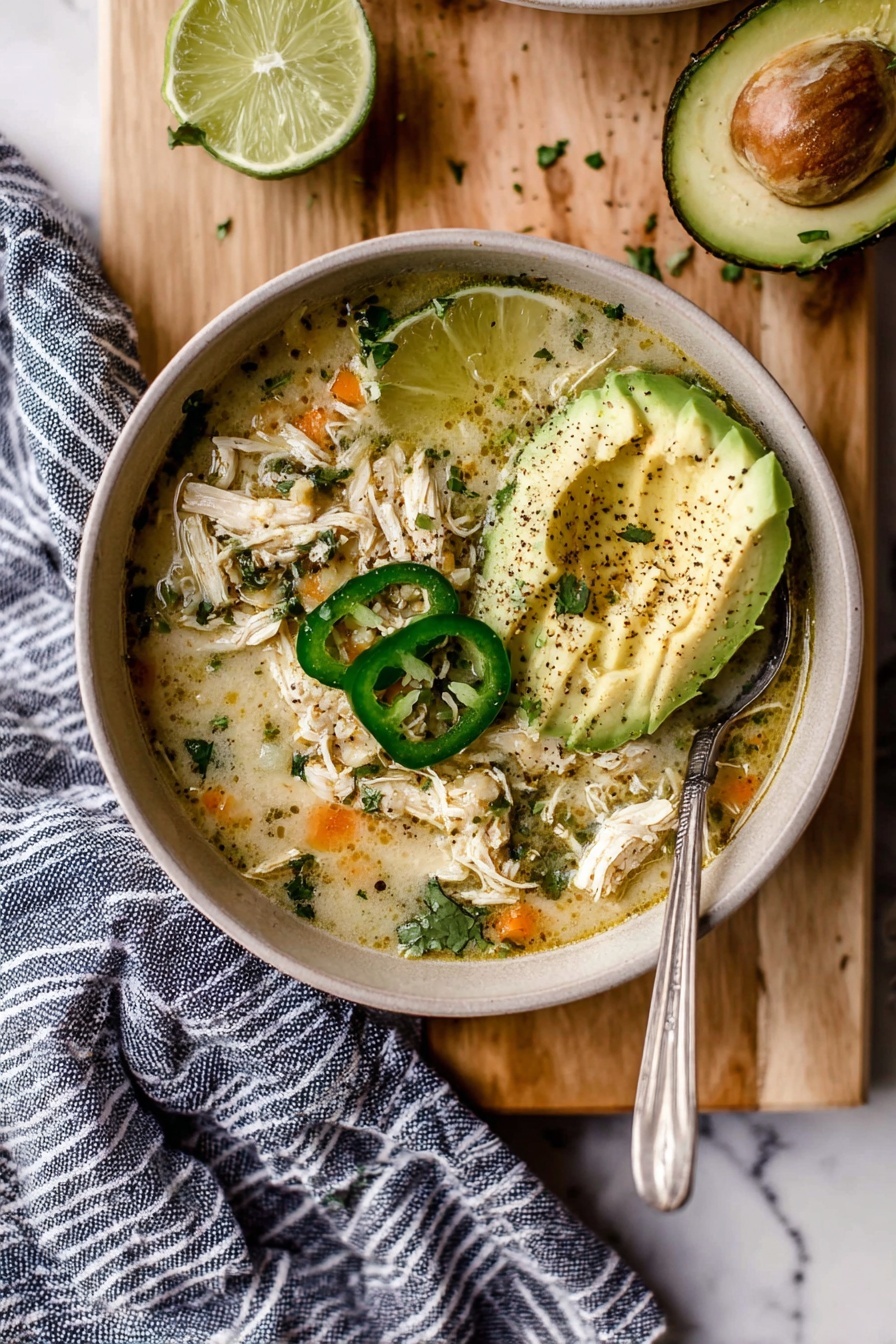 A bowl of soup with shredded chicken mixed in a light, creamy broth, showing bits of herbs and small orange pieces. On top, there are three green jalapeño slices placed near the center, and a pale green avocado fan with black pepper sprinkled over it to the right. A lime wedge sits just above the avocado, slightly pressed. A spoon is placed on the right side inside the bowl. The bowl is set on a light wooden board with a cut lime half to the left and a striped blue and white cloth draping off the right side. The background is a white marbled texture. photo taken with an iphone --ar 2:3 --v 7