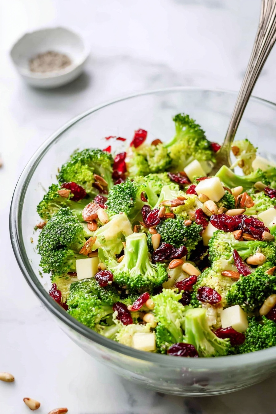 A clear glass bowl filled with fresh broccoli salad sits on a white marbled surface. The salad has bright green broccoli florets mixed with small cubes of pale yellow cheese, deep red dried cranberries, and glossy pieces of nuts, likely almonds, scattered evenly throughout. There are also light brown sunflower seeds sprinkled atop the salad. A silver fork is resting inside the bowl, angled towards the right side of the image. In the background, a small white dish containing some coarse salt and pepper is slightly blurred. A woman's hand is not visible in this image. photo taken with an iphone --ar 2:3 --v 7