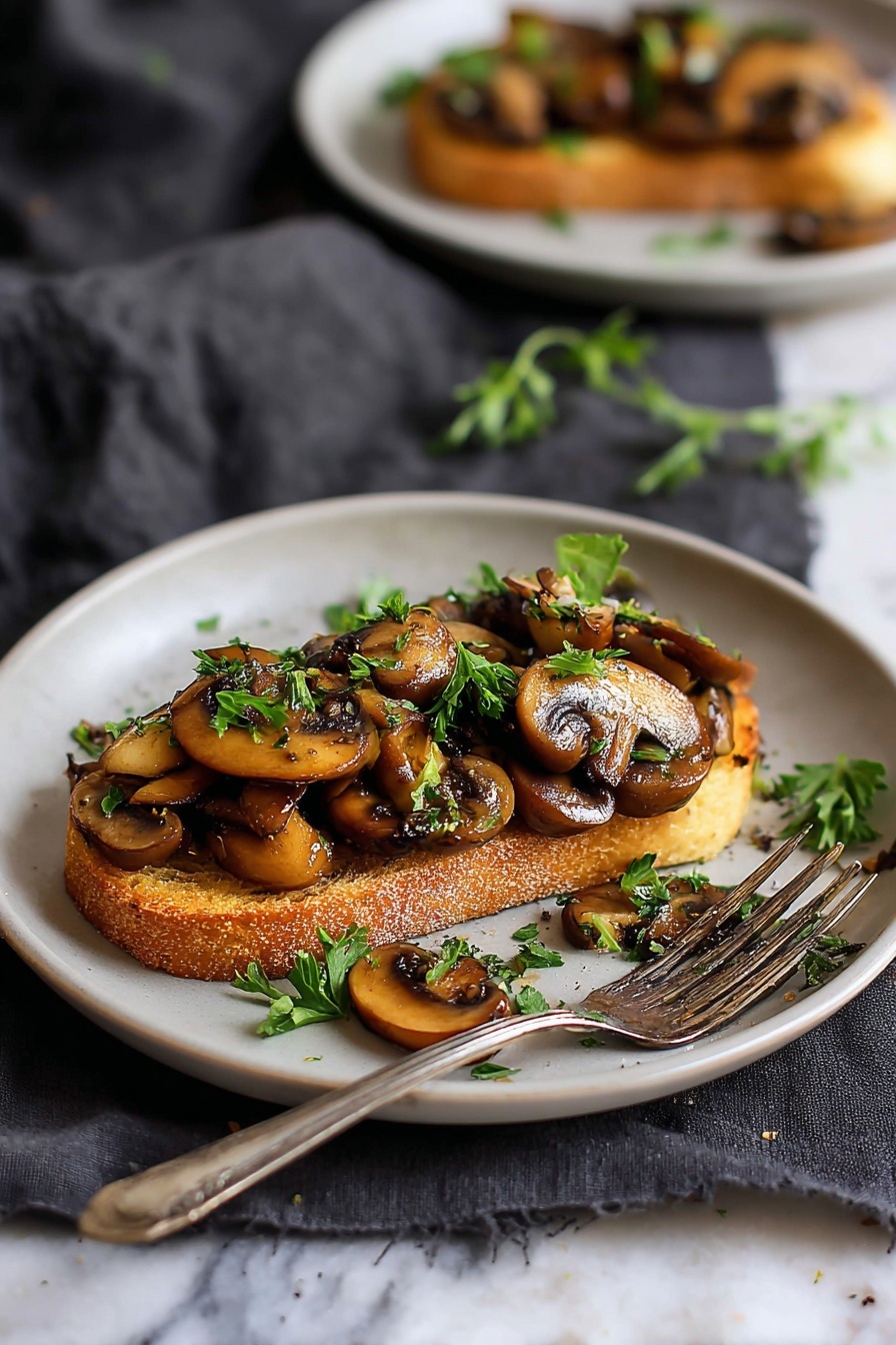 The image shows a white plate with one slice of golden brown toasted bread as the base layer, topped with sautéed mushrooms that are light to dark brown, some with a slight shine, scattered evenly on top. Bright green chopped herbs are sprinkled on the mushrooms for contrast. A silver fork rests on the right side of the plate, which sits on a gray cloth over a white marbled surface. Some mushroom pieces and herbs are scattered lightly around the plate and surface. Photo taken with an iphone --ar 2:3 --v 7