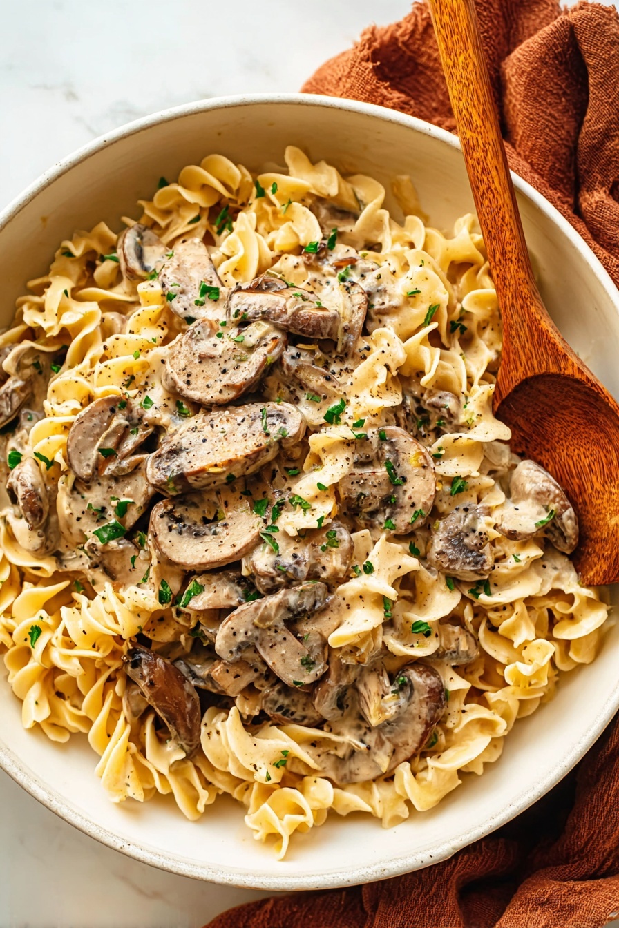 A white bowl holds a creamy mushroom noodle dish with two main layers: a bed of pale yellow, curly egg noodles forming the base and sauce-soaked sliced brown mushrooms spread evenly over them. The creamy sauce is light beige with specks of black pepper, coating both the noodles and mushrooms. Small green herb bits scatter on top, adding a fresh contrast. On the right side of the bowl, a wooden spoon with a warm brown tone dips slightly into the noodles and mushrooms. The bowl rests on a soft, rust-colored cloth, all set against a white marbled surface. Photo taken with an iphone --ar 2:3 --v 7