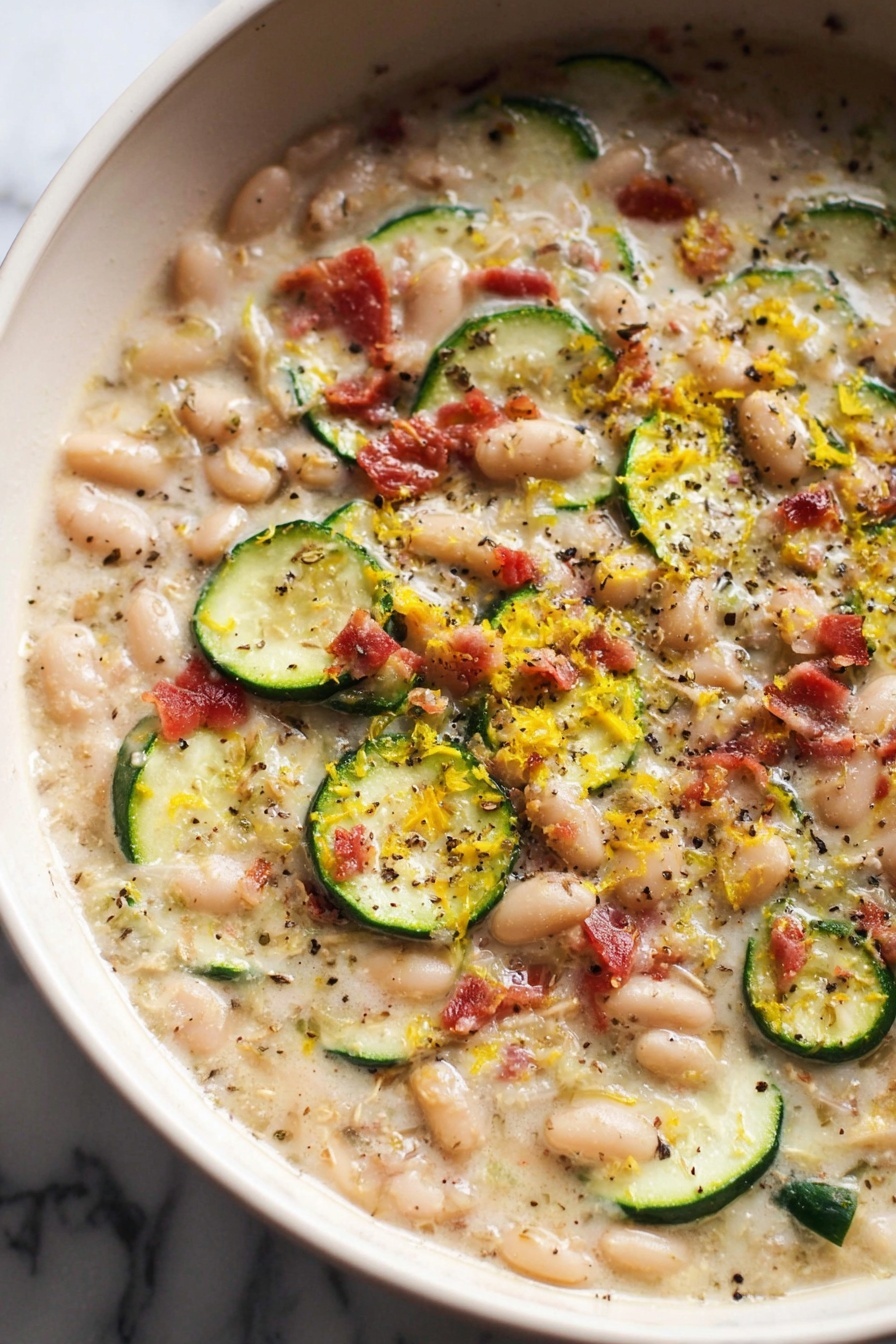 This dish is in a white bowl with a smooth, creamy base filled with many small white beans spread around. It has thin, round slices of green zucchini mixed in, along with small pieces of red bacon scattered on top. The surface is sprinkled with black pepper and yellow zest, giving a fresh and textured look. The creamy liquid looks rich and slightly shiny, holding all these colorful ingredients together. The background is a white marbled surface. photo taken with an iphone --ar 2:3 --v 7