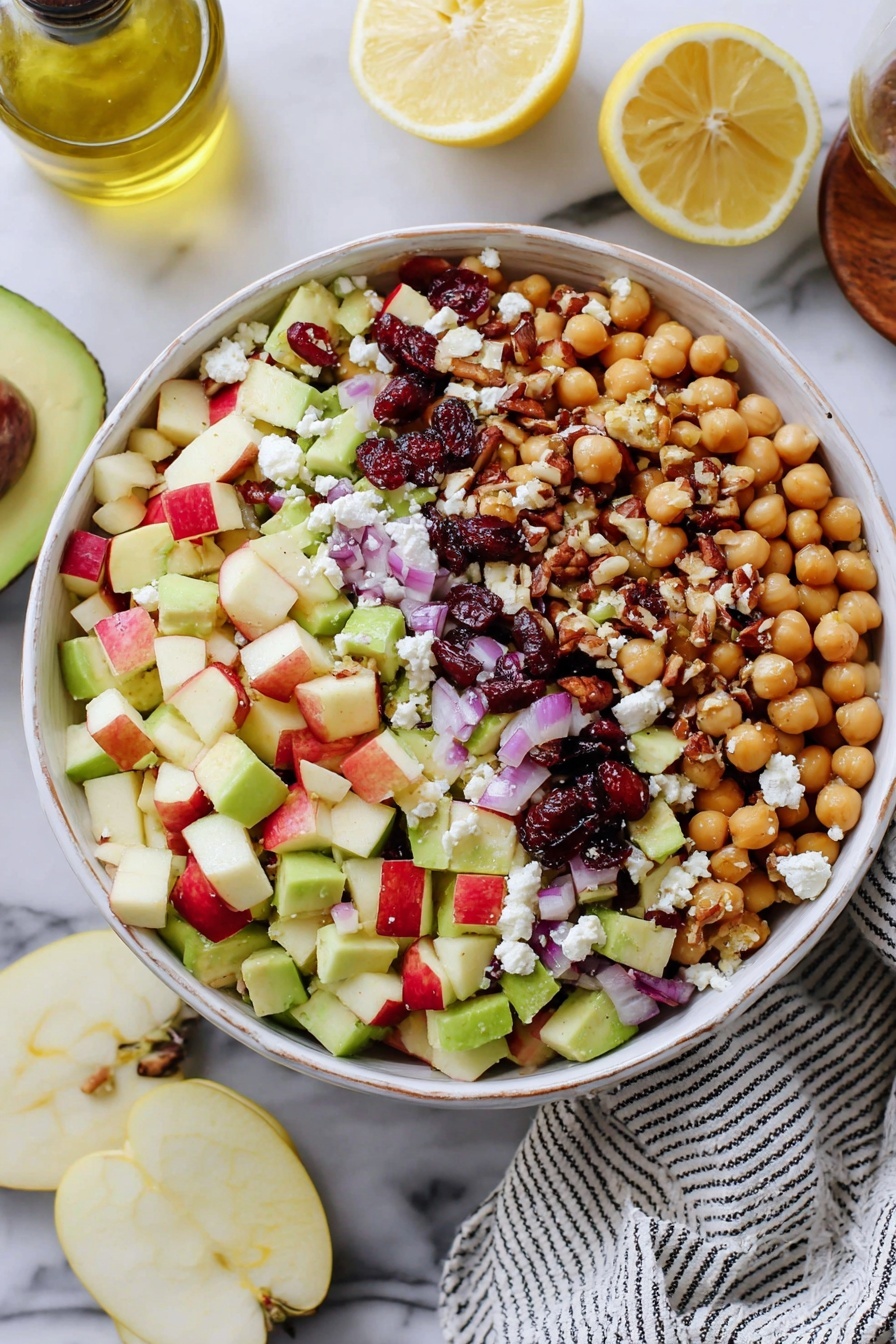A white bowl filled with a colorful salad sits on a white marbled surface. The salad has four main layers: a base layer of round, beige chickpeas, a second layer of diced red and green apples with light green avocado chunks, a third layer of small pieces of red onion and dark red dried cranberries, and a top layer of white crumbled feta cheese mixed with brown chopped nuts. On the side of the bowl, there are three small lemon wedges placed within the salad. Around the bowl, you can see half a lemon, a glass bottle of olive oil, an apple half, a few slices of avocado, and a white and black striped cloth partially visible. The photo taken with an iphone --ar 2:3 --v 7