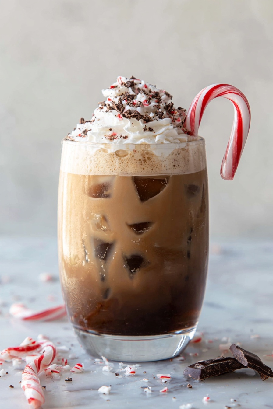 A clear glass holds a three-layered iced drink on a white marbled surface. The bottom layer is dark brown with bits of chocolate and candy cane pieces visible inside. The middle layer is a lighter brown with ice cubes floating in it, giving a cool and fresh look. The top layer is creamy white whipped cream with dark chocolate crumbs sprinkled over it. A white and red striped candy cane is hooked on the side of the glass. Pieces of broken candy cane and chocolate are scattered around the base of the glass. photo taken with an iphone --ar 2:3 --v 7