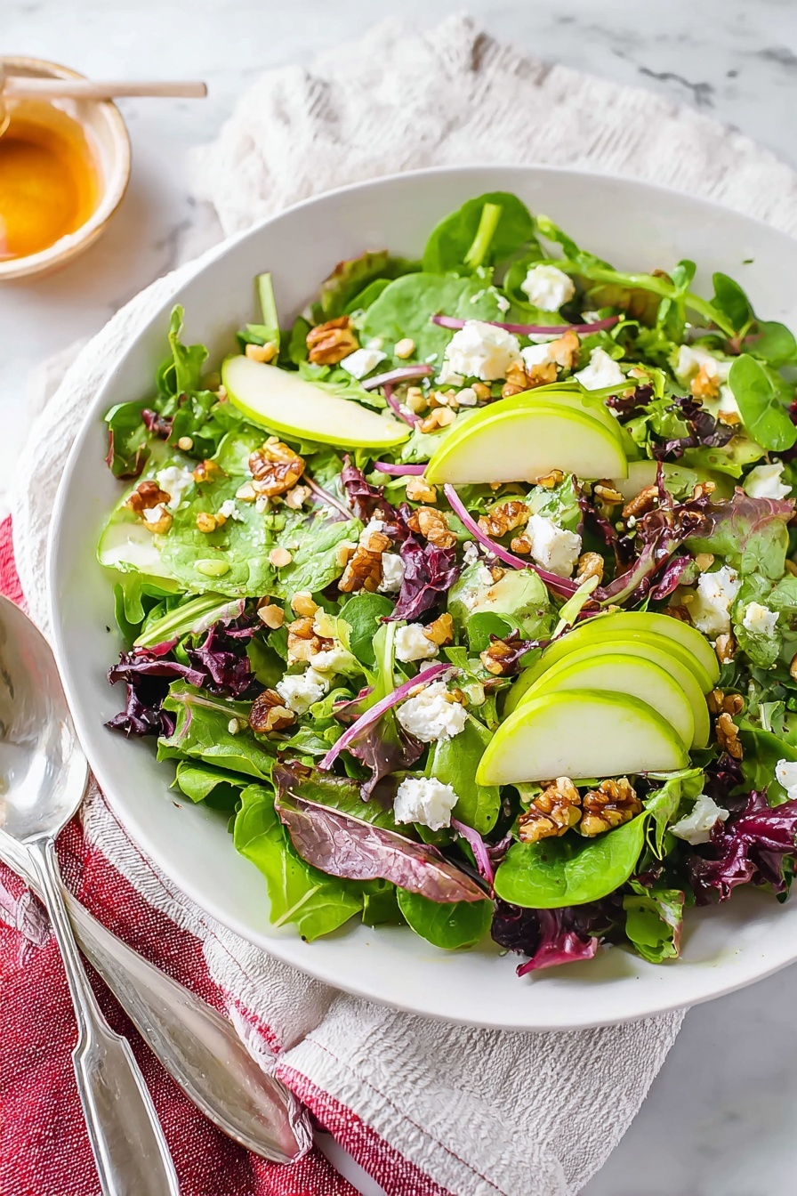 The dish is a fresh salad served on a white plate, placed on a white marbled surface. The salad has three main layers: the base is bright green leafy spinach and mixed greens, the middle layer contains light green slices of apple and small pieces of walnuts that add texture, and the top layer includes small white chunks of cheese and bits of red onion scattered evenly. There are also dark red dried cranberries spread throughout the salad, adding color contrast. A silver fork and spoon rest on the left side of the plate, lightly touching the salad. In the background, a wooden cutting board with a green apple and a knife are blurred but visible. photo taken with an iphone --ar 2:3 --v 7