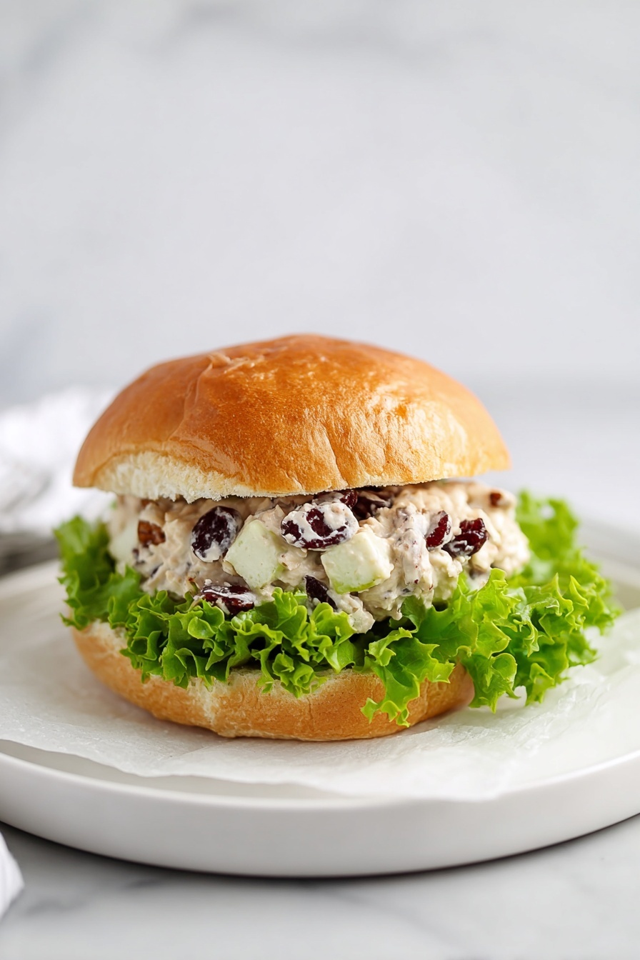 A sandwich with a golden brown bun that looks soft and slightly cracked on the top layer. Inside, there is a layer of fresh, frilly green lettuce at the bottom. Above the lettuce, there is a thick, creamy mixture with visible small pieces of white apple, dark dried cranberries, and brown pecans, all mixed in a white sauce. The sandwich is placed on a white plate with a sheet of white paper underneath on a white marbled surface. photo taken with an iphone --ar 2:3 --v 7