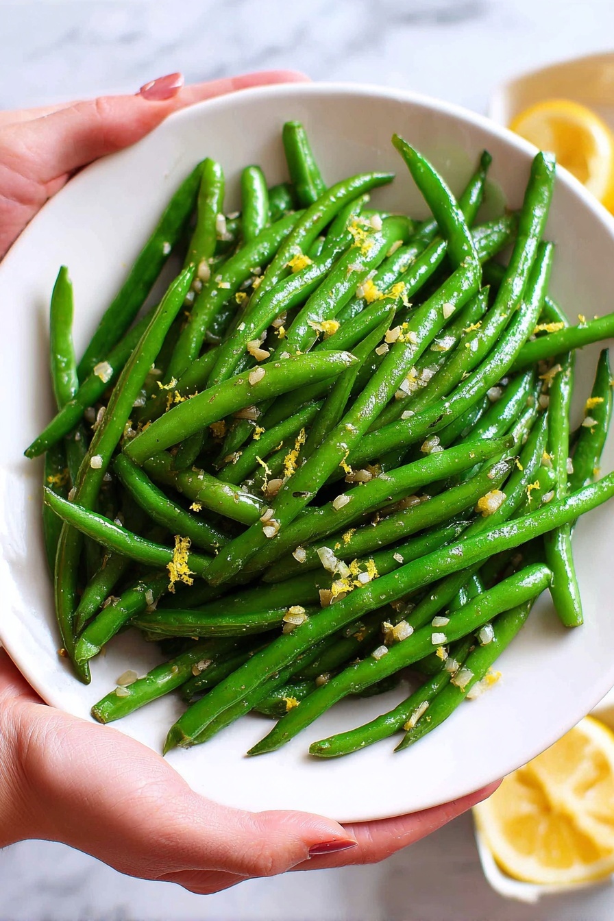 A white round plate full of bright green cooked green beans is shown being held by a woman's hand on each side. The green beans are spread in one thick layer across the plate, mixed with small bits of minced garlic and tiny yellow lemon zest pieces scattered on top. The background is a white marbled surface, with a small part of a container with lemon wedges blurred out in the corner. Photo taken with an iphone --ar 2:3 --v 7