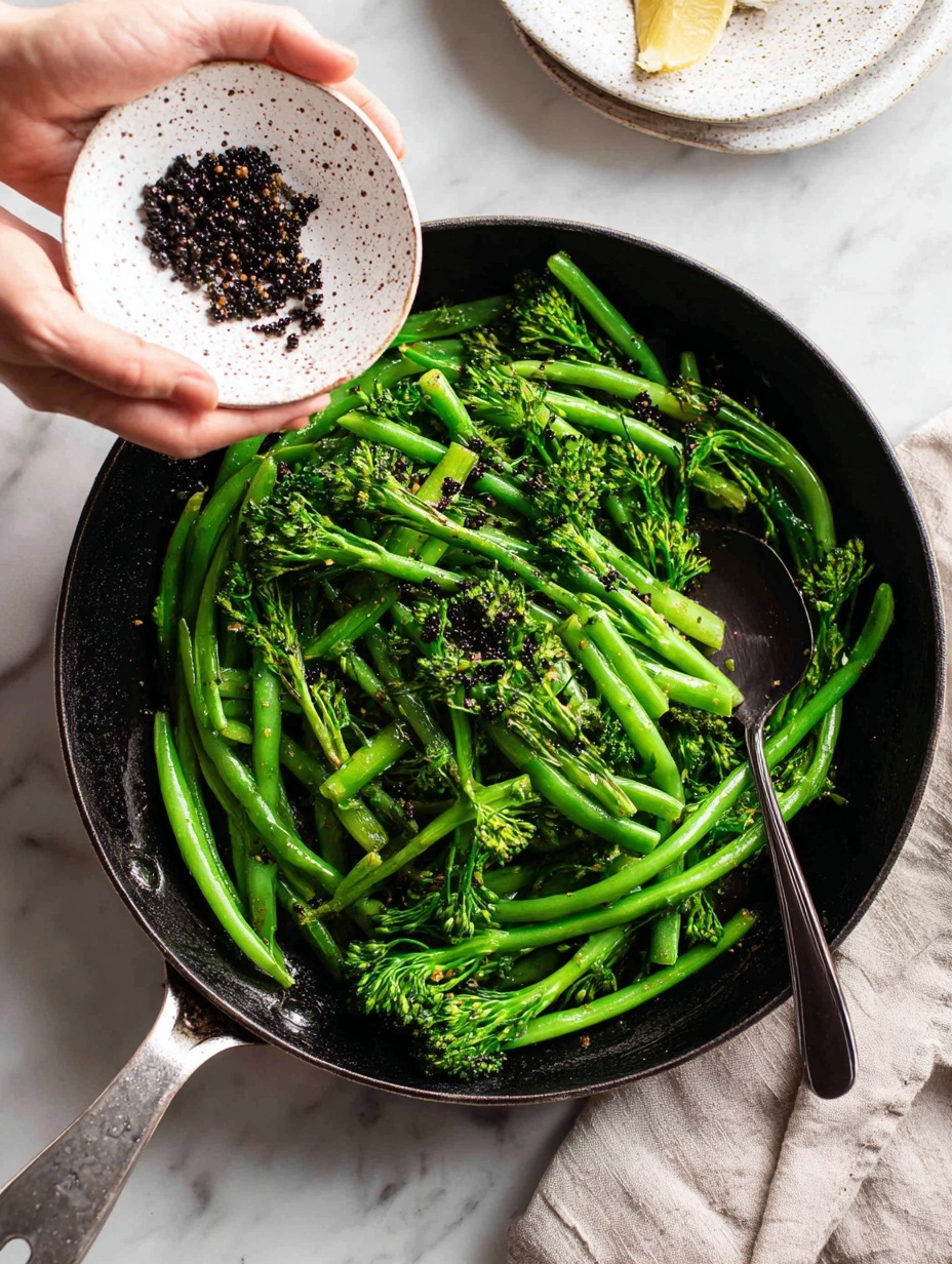 A skillet filled with bright green long green beans and broccolini, showing fresh and slightly glossy textures from cooking. Above the skillet, a woman's hand is holding a white speckled small plate pouring small dark lentils or seeds into the skillet. A dark spoon with the brand name
