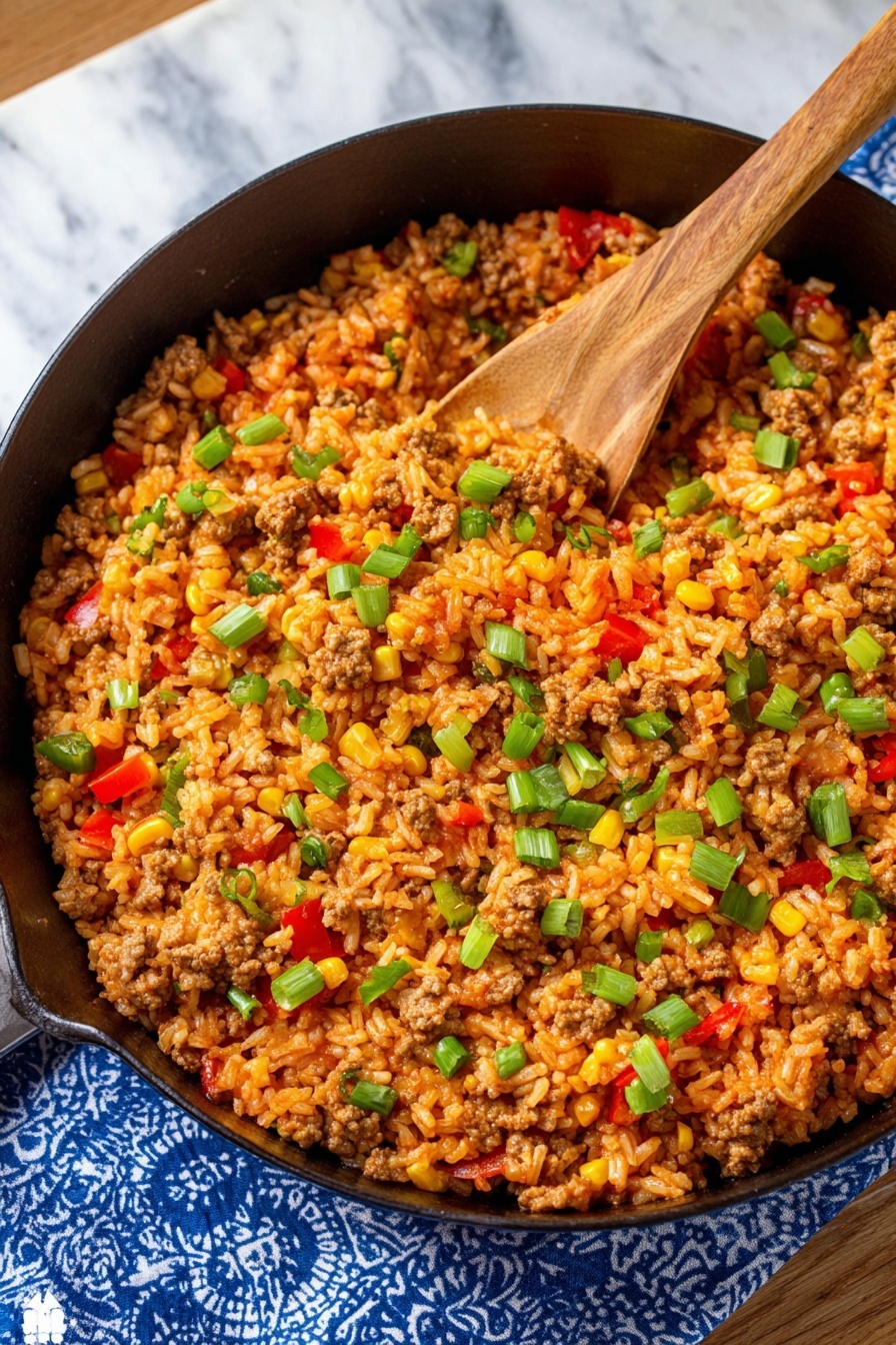The image shows a black cast iron pan filled with a single layer of cooked rice mixed with ground meat, corn, diced red and green bell peppers, and small pieces of green onions scattered on top. The rice has a reddish-orange color, likely from a sauce, and is mixed evenly with the other ingredients. A wooden spoon rests inside the pan, partly submerged in the rice mixture. The pan is set on a white marbled surface with a blue and white patterned cloth partially visible underneath the pan. Photo taken with an iphone --ar 2:3 --v 7