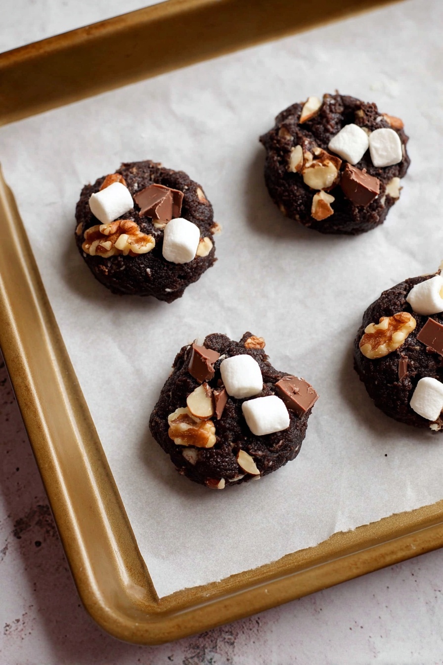 Four small, round dark chocolate cookie dough balls sit spaced apart on a baking tray lined with white parchment paper. Each ball is topped with chunks of dark brown chocolate, small white marshmallows, and whole light brown almonds, creating a textured, chunky look. The dark cookie dough base features bits of walnuts visible inside, adding a mix of rough texture and earthy color. The tray rests on a surface with a faint white marbled texture. photo taken with an iphone --ar 2:3 --v 7
