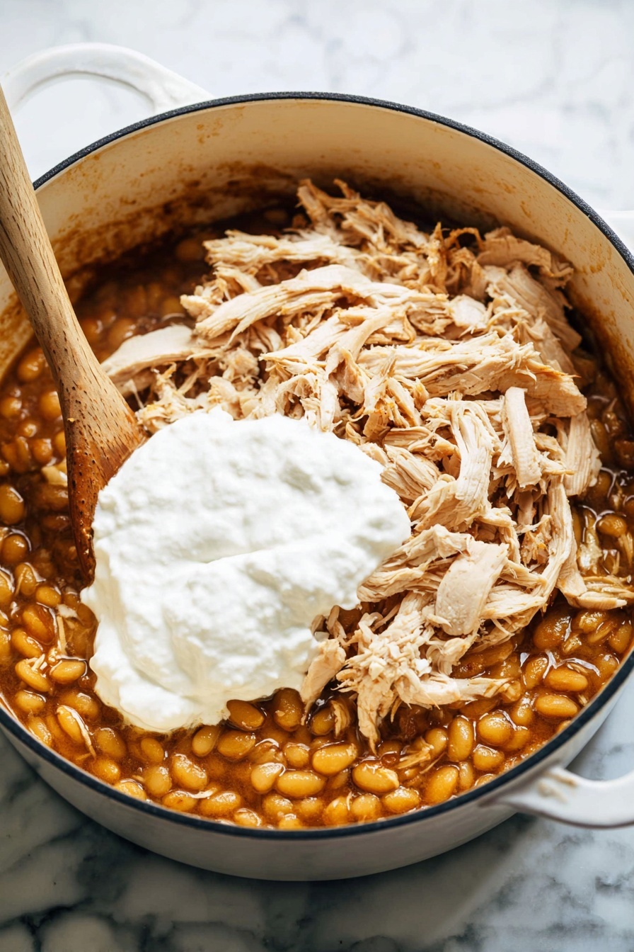 A white pot is shown on a white marbled surface, filled with three visible layers of food. The bottom layer consists of yellowish beans in a brownish sauce, covering the whole base. On top of this is a layer of shredded light brown chicken pieces, spread unevenly across. The top layer is a dollop of thick white cream sitting in the center over the chicken. A wooden spoon sticks into the pot from the left side. Photo taken with an iphone --ar 2:3 --v 7