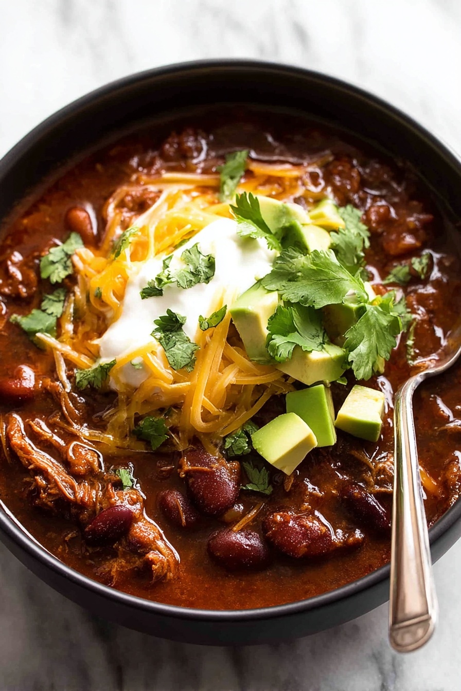 A black bowl filled with a thick stew made of brown shredded meat and red beans in a rich reddish-brown sauce. On top, there is a layer of melted yellow cheese scattered unevenly. Above the cheese, a dollop of white sour cream is placed in the center. Around the sour cream, small chunks of bright green avocado and pieces of fresh green cilantro are scattered. A silver spoon is resting inside the bowl on the right side with its handle leaning out. The bowl sits on a surface with a white marbled texture. Photo taken with an iphone --ar 2:3 --v 7