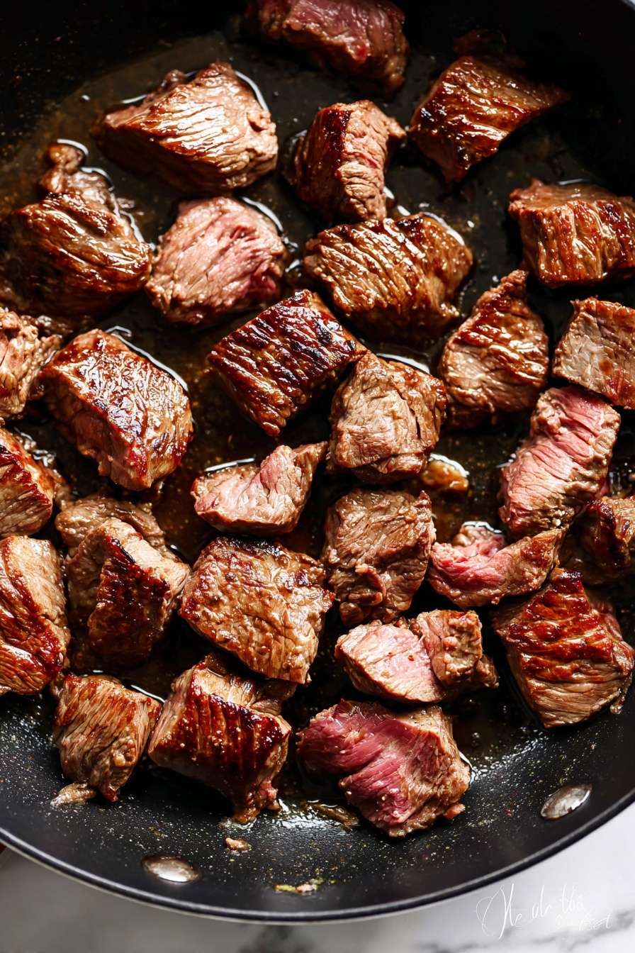 The image shows many small pieces of cooked beef in a black pan. The beef pieces are irregular in shape with a browned outside and some pinkish spots inside, giving a rich texture. The beef chunks are spread across the pan with some shiny spots of oil around them. The pan surface is dark and slightly greasy with reflections from the cooked meat. photo taken with an iphone --ar 2:3 --v 7