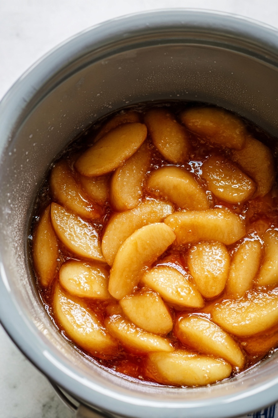 The image shows light golden apple slices in a thick, shiny, warm amber sauce inside a gray cooking pot with a smooth inner surface and water droplets on the sides. The apple slices are soft and coated well in the sauce, which looks like it has a syrupy texture, filling the bottom of the pot evenly. The inner sides of the pot show condensation, adding to the sense of warmth and cooking in progress. The scene is set on a white marbled texture surface. photo taken with an iphone --ar 2:3 --v 7